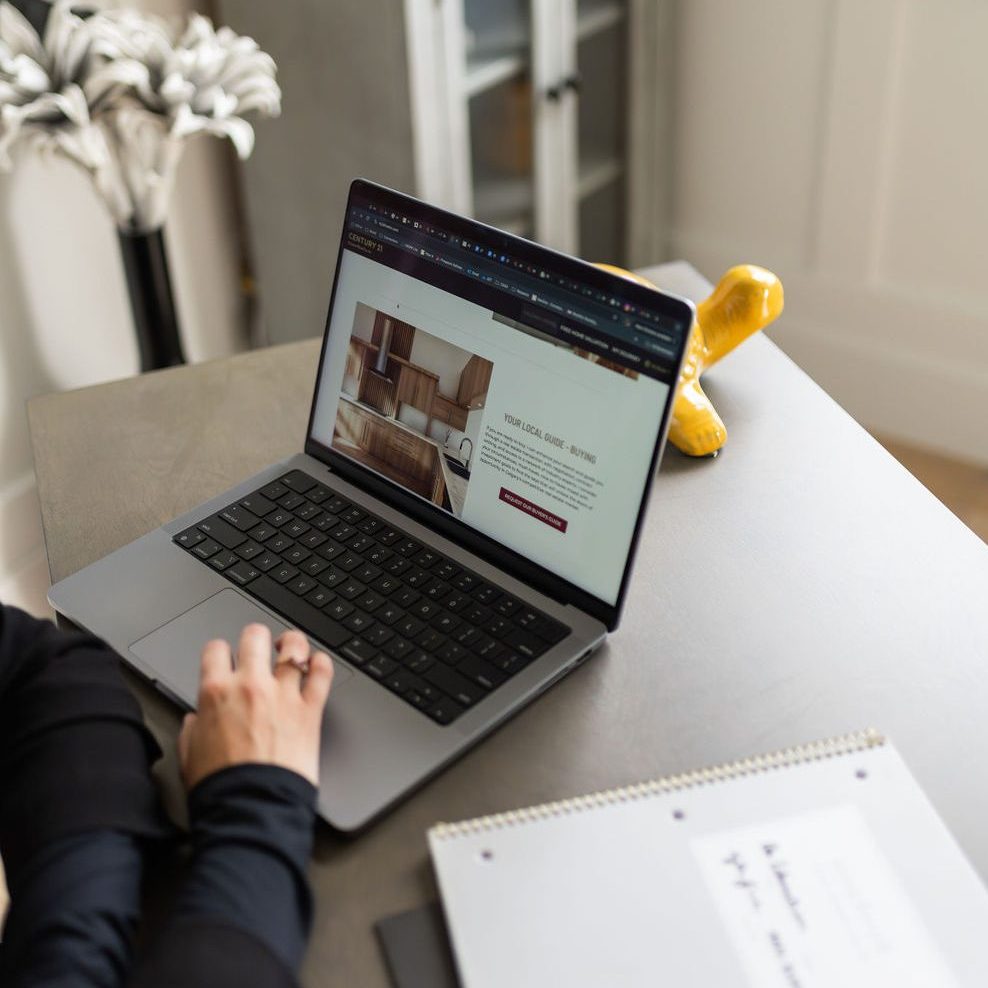 Woman is searching a realtors website, wearing all black with arms crossed and a notebook and folder on the side of the wooden desk