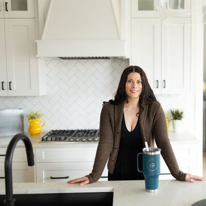 Woman standing in a light and bright kitchen with a teal yeti mug in front of her, smiling, leaning against the stone counter top.